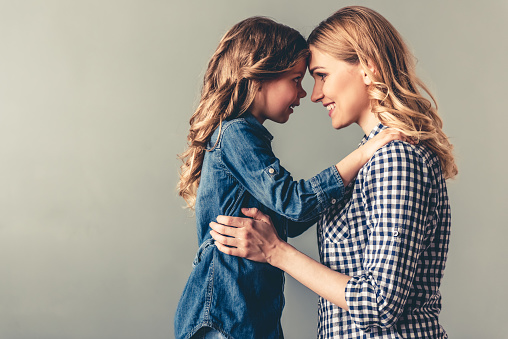 Cute little girl and her beautiful young mom are touching their foreheads, looking at each other and smiling, on gray background