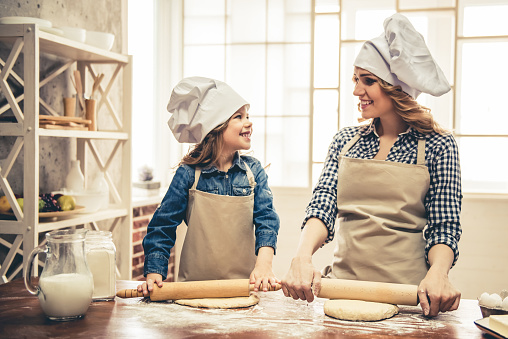 Cute little girl and her beautiful mom in aprons and chef hats are smiling while flattening the dough using a rolling pin in the kitchen