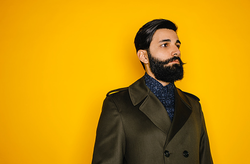 Portrait of bearded serious brunette middle eastern young male hipster standing over a yellow background, side view