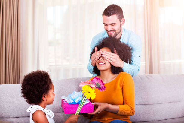 father holding gift and flowers, daughter covering mothers eyes - men african descent giving flower imagens e fotografias de stock
