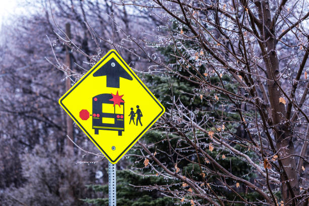 A brand new, bright yellow, red, and black diamond-shaped road sign alerting drivers to a school bus stop ahead where school children will be walking, waiting for, boarding, and/or exiting education transportation school buses. The symbols on the sign indicate that when any school vehicle is stopped on any roadway - with its side warning stop signs extended and/or its roof level red warning lights flashing - New York State traffic law requires all other vehicle drivers approaching from any direction to stop, and not pass the school vehicle until extended stop signs are fully retracted and all warning lights are no longer flashing. This sign is located in a rural western New York State suburban area near Rochester, NY, USA. Photo taken in December during a dangerous, freezing sleet storm with heavy ice accumulations.