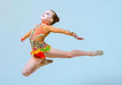 Beautiful elementary age girl dressed in a gymnastic leotard is showing a rhythmic gymnastics elements. The girl is jumping sideway to the camera. She is in flight and looking forward. Studio shooting on light blue background
