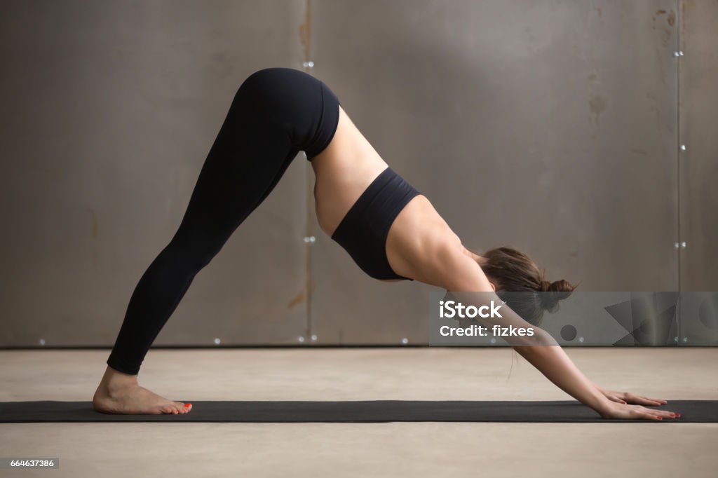 Young attractive woman in adho mukha svanasana pose, grey studio Young attractive yogi woman practicing yoga, standing in adho mukha svanasana exercise, Downward facing dog pose, working out, wearing black sportswear, cool urban style, full length, grey studio Adult Stock Photo