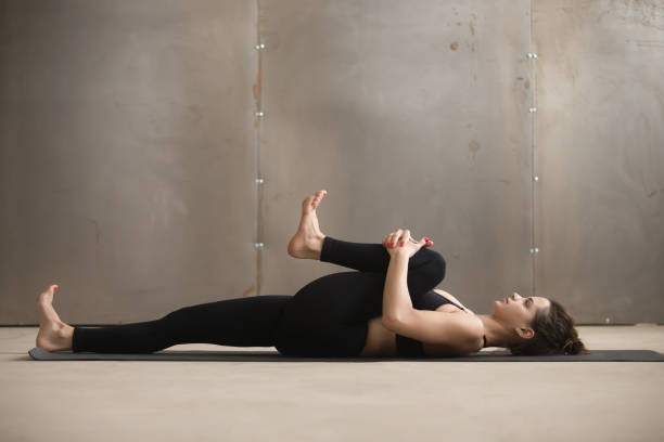 Young woman in Knees to Chest, Apanasana pose, grey studio Young attractive sporty woman practicing yoga, stretching in Knees to Chest, Apanasana exercise, working out, wearing black sportswear, cool urban style, grey studio background, full length, side view shoulder stand stock pictures, royalty-free photos & images