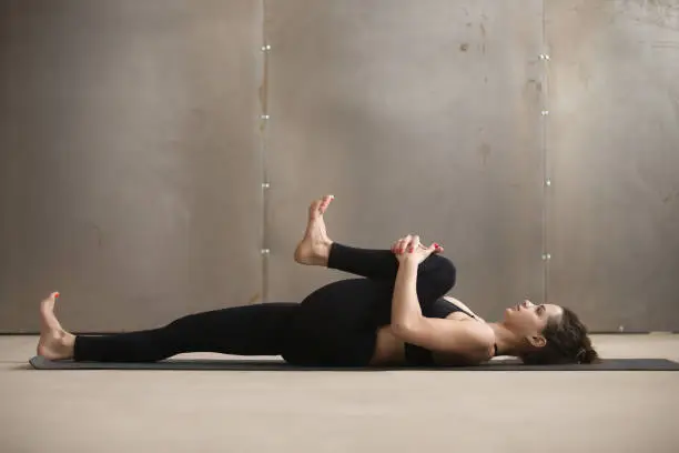 Young attractive sporty woman practicing yoga, stretching in Knees to Chest, Apanasana exercise, working out, wearing black sportswear, cool urban style, grey studio background, full length, side view