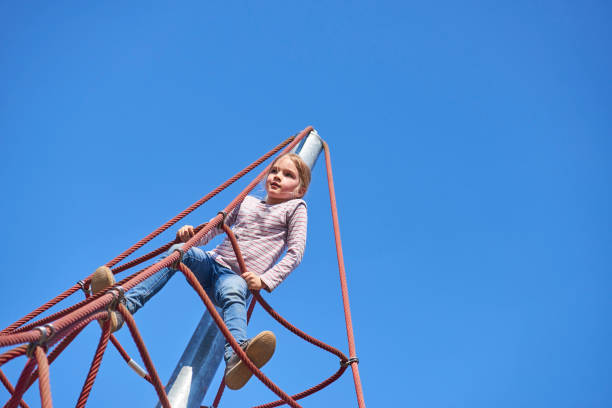 A cheerful child triumphantly raises their arm atop a colorful rope structure in a sunny park, surrounded by vibrant blue skies and greenery, symbolizing joy, achievement, and youthful energy.