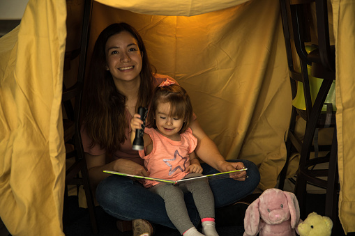Happy young mom sitting with her pretty child reading a book holding a flashlight inside a tent in the living room