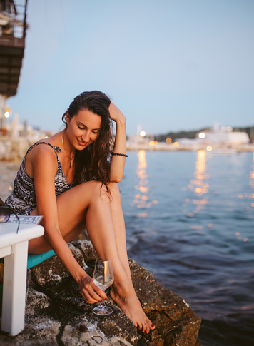 Portrait of a young woman, sitting by the beach in an outdoors cafe in the Croatian seaside, Mediterranean. She is enjoying watching the sunset, having a glass of wine in the dusk by the sea.
