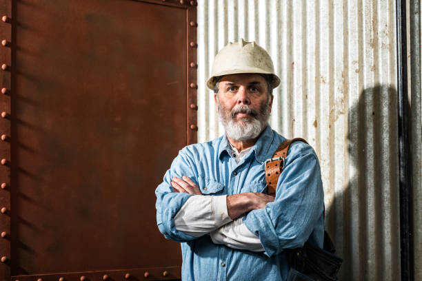 Rugged looking Middle-aged construction worker on job site A portrait of a rugged looking middle-aged Caucasian male construction worker with a gray beard, wearing an aluminum hard hat and has a leather tool belt over his shoulder, looking at the camera with his arms crossed, with an old rusted steel plate and corrugated steel background. He is also wearing a blue denim shirt with a off white Henley underneath. construction worker photos stock pictures, royalty-free photos & images