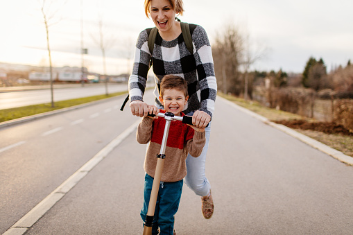 Photo of a cheerful little boy riding on the same scooter with his mom.