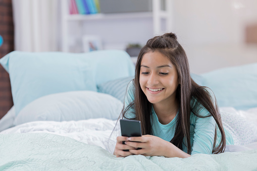 Happy Hispanic tween girl uses a smart phone while in her room.