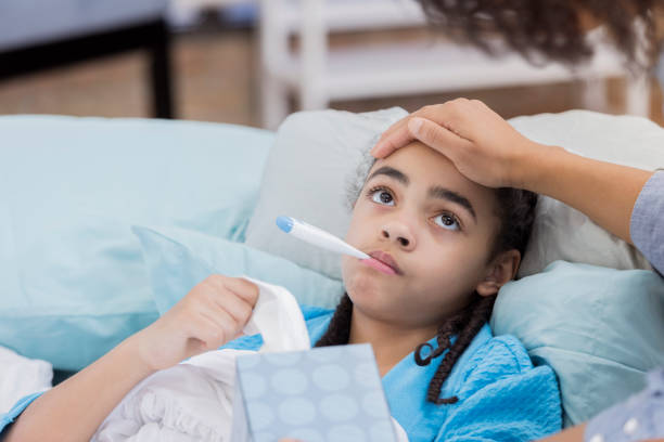 African American mother checks her daughter's temperature. The girl has a thermometer in her mouth and is holding a box of tissues.