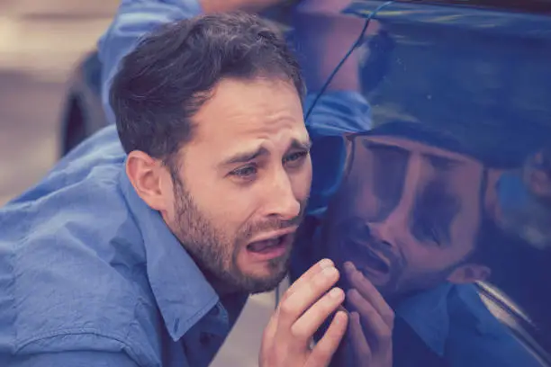 Photo of Frustrated upset young man looking at scratches and dents on his car outdoors