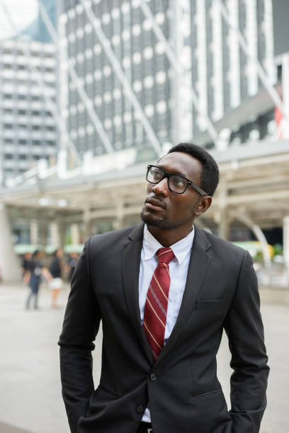 istock Young African businessman thinking while looking up with eyeglasses in front of modern building at Bangkok city 664871556