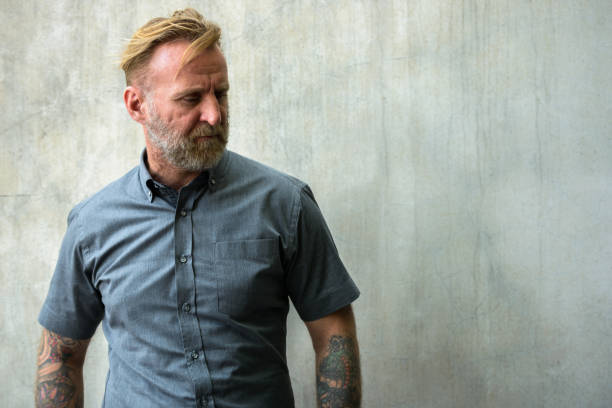 istock Mature bearded man with hand tattoos thinking while looking down against urban concrete wall 664871676