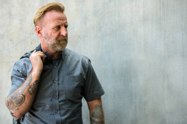 istock Mature bearded man with hand tattoos thinking while holding jacket over the shoulder against urban concrete wall 664871798
