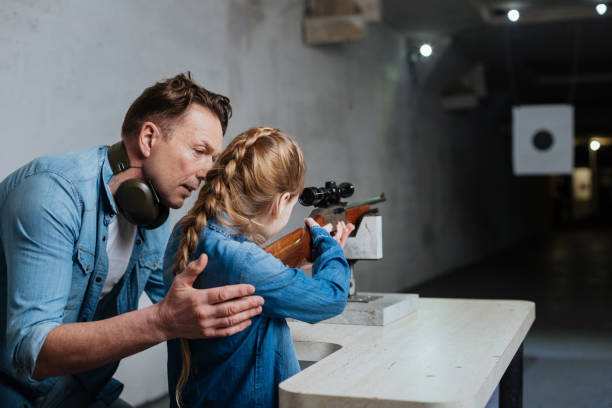 Handsome caring father teaching his daughter Time together. Handsome nice caring father standing near his daughter and supporting her while teaching her how to shoot target shooting stock pictures, royalty-free photos & images