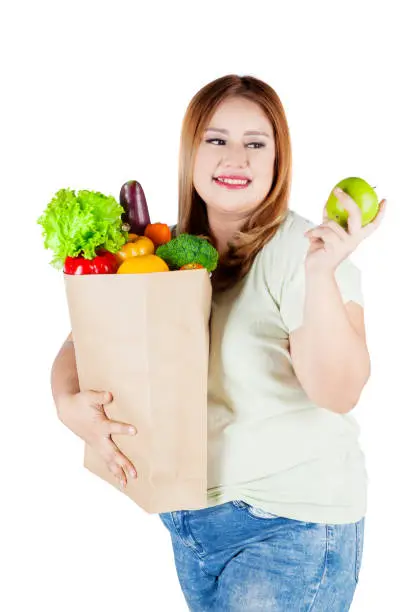 Portrait of an overweight woman with blonde hair, holding an apple fruit and a shopping bag full of healthy foods