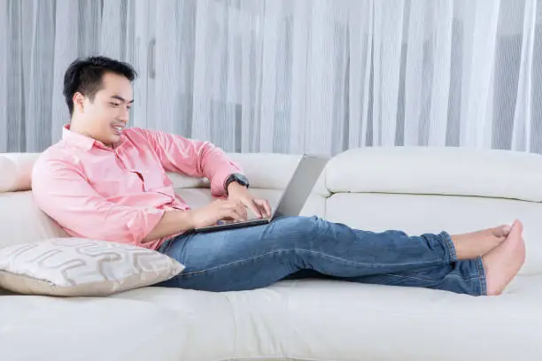 Portrait of young male lying and relaxing on the couch while using a laptop in the living room