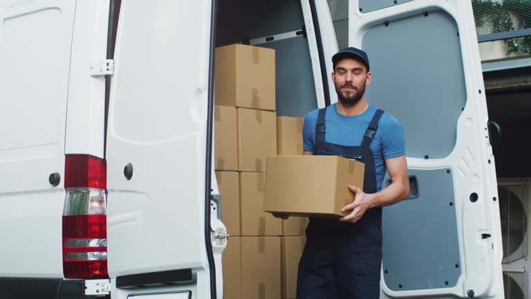 Young and Smiling Delivery Service Man Takes Cardboard Box from his Cargo Van and Walks towards Delivery Destination.