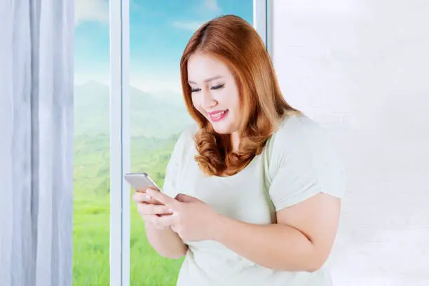 Picture of happy blonde woman with overweight body, texting on her smartphone while standing near the window