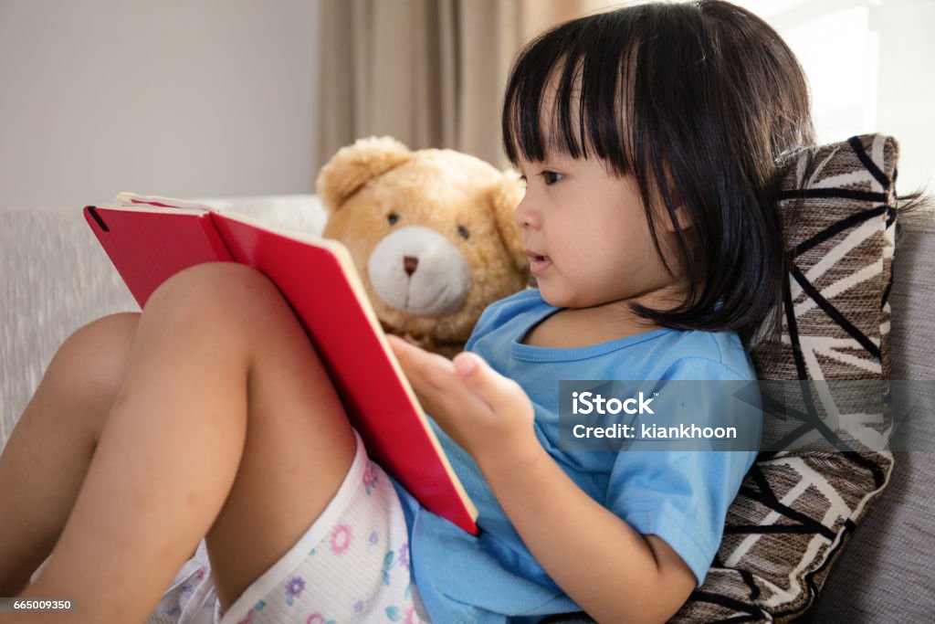 Asian Chinese little girl reading book with teddy bear Asian Chinese little girl reading book with teddy bear on the couch in the living room at home. Child Stock Photo