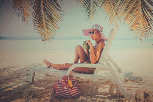 Cute blonde girl enjoying on her vacation. She is laying down in easy chair drinking from her coconut glass with straw. On her left she has her bag and sun lotion. She is laying down under coconut tree which provides her cover and shadow from burning sun. It's beautiful and sunny day outside, and she is really enjoying on the beach. She is wearing two piece swimsuit, sunglasses and pink hat on her head.