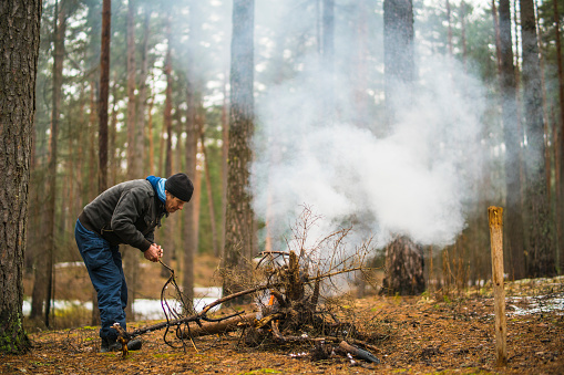 Active senior, 70-years-old, caucasian white man make the fier in the forest for heat and fun at the rest during the travel. Early spring, cold sunny weather, Belarus, eastern Europe