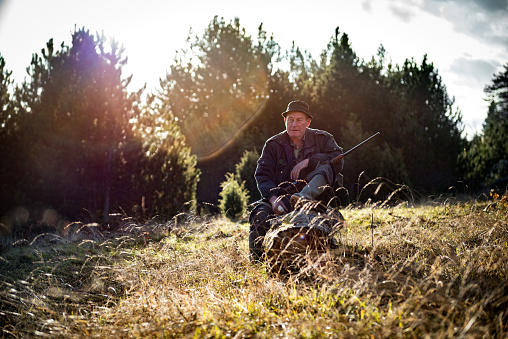 Portrait of a hunter in the forest while sitting on the tree log and holding a rifle.