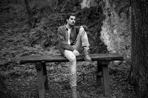 Beautiful young man sitting on the wooden table in the forest and smoking cigarette