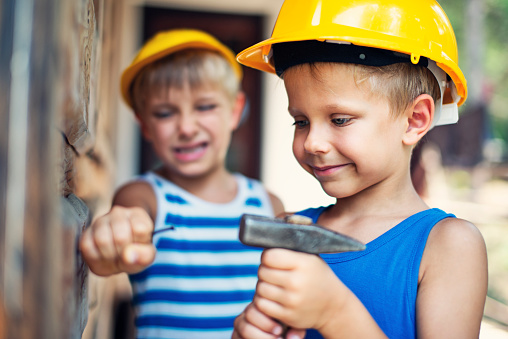 Little boys dressed up as manual workers aged 5 are hammering a nail to the wooden cabin wall.  Little guys are doing the task together even though one of them is taking all the risk on him.\n