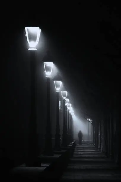 person walking on dark street illuminated with streetlamps. Black and white