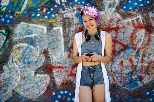 Smiling young hipster girl using smart phone against graffiti wall