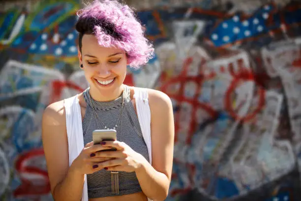 Photo of Smiling young hipster girl using smart phone against graffiti wall