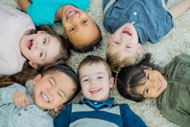Big Smiles A multi-ethnic group group of young children lay down in a circle on a living room carpet with their heads together all brightly smiling at the camera. mixed racial group of friendly children laughing together stock pictures, royalty-free photos & images
