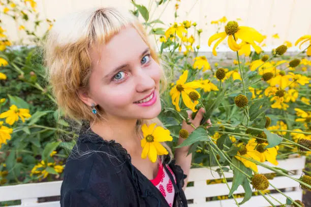 Closeup of young woman smiling by tall sunflower yellow daisy flowers on bench