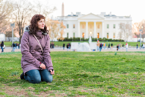 Young woman sitting in grass in park in front of white house at sunset