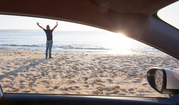 istock Man stretches outside car on beach, by sea shore 665267753