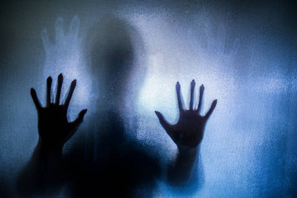 Spooky silhouette of woman with hands pressed against glass window Colour backlit image of the silhouette of a woman with her hands pressed against a glass window. The silhouette is distorted, and the arms elongated, giving an alien-like quality. The image is sinister and foreboding, with an element of horror. It is as if the 'woman' is trying to escape from behind the glass. Horizontal image with copy space. people trafficking stock pictures, royalty-free photos & images