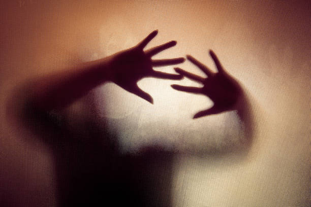 Spooky silhouette of woman with hands pressed against glass window Colour backlit image of the silhouette of a woman with her hands pressed against a glass window. The silhouette is distorted, and the arms elongated, giving an alien-like quality. The image is sinister and foreboding, with an element of horror. It is as if the 'woman' is trying to escape from behind the glass. Horizontal image with copy space. sexual assault stock pictures, royalty-free photos & images
