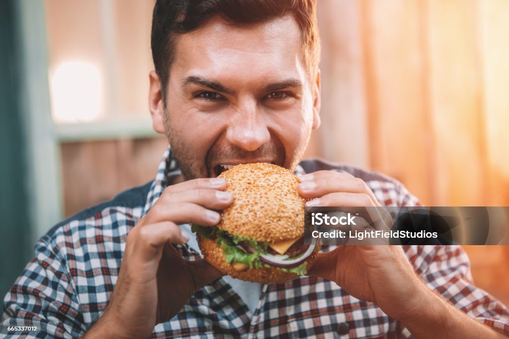 Hombre joven de morder la hamburguesa sabrosa fresca y mirando a cámara - Foto de stock de Hamburguesa vegetariana libre de derechos