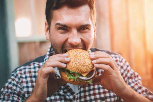 istock Young man biting fresh tasty hamburger and looking at camera 665337012