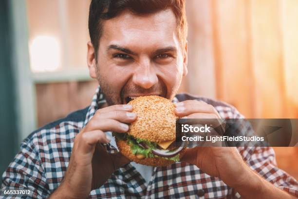 Young Man Biting Fresh Tasty Hamburger And Looking At Camera Stock Photo - Download Image Now