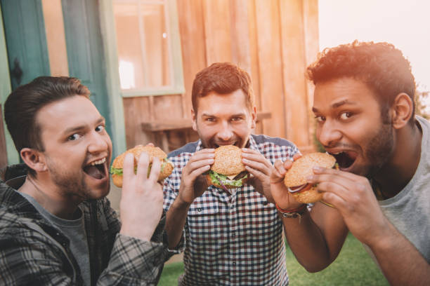istock Three smiling young men eating fresh hamburgers and looking at camera 665337424
