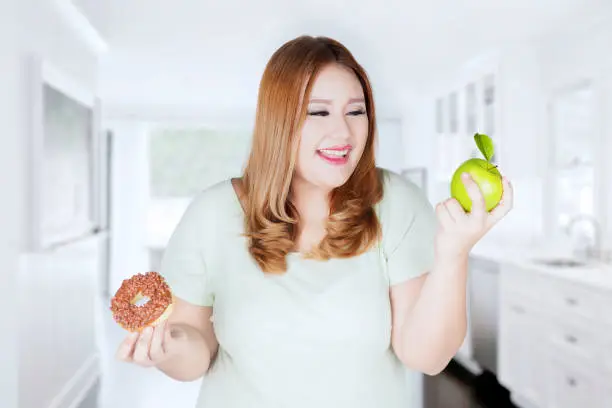 Obese woman with blonde hair, choosing between apple fruit or donut in the kitchen