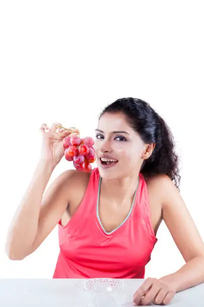 Young Indian woman holding a bunch of ripe grapes while looking at the camera and sitting in the studio