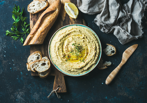 Homemade hummus with lemon, herbs and freshly baked baguette on rustic wooden serving board over dark blue plywood background, top view, horizontal composition, Vegetarian food concept