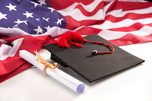Close-up view of graduation mortarboard, diploma and us flag on white, education concept