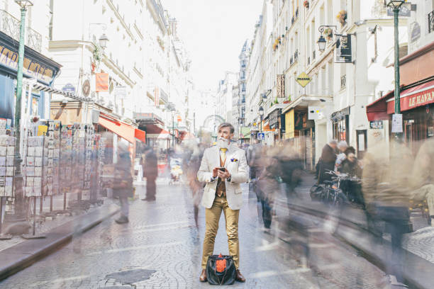 Businessman holding mobile phone while standing amidst crowd moving on street. Full length of professional is on business tour in city. Thoughtful professional is looking away.