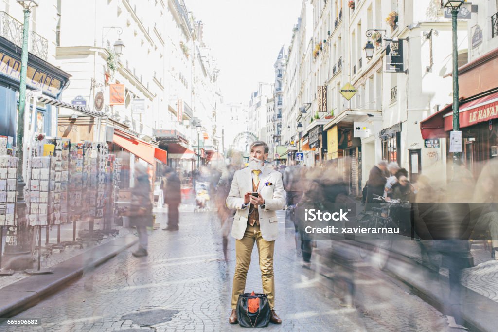 Businessman standing amidst crowd moving on street Businessman holding mobile phone while standing amidst crowd moving on street. Full length of professional is on business tour in city. Thoughtful professional is looking away. Crowd of People Stock Photo